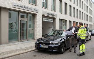 Ein Architekt im Anzug steht neben einem Mitarbeiter eines Abschleppdiensts und zeigt auf ein Auto, das eine Einfahrt blockiert.